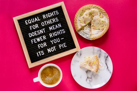 A pie sits on a pink table with one slice seperate on a plate next to a coffee. There is a board with the words 'Equal rights for others doesn't mean fewer rights for you - it's not pie'