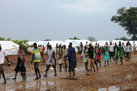 South sudan refugees in Ethiopia walk through a flooded dirt road infront of rows of tents