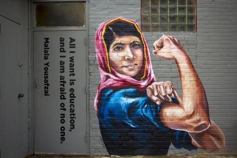 A painted mural of Malala Yousafzai on a brick wall. She wears a headscarf and is flexing her bicef in a display of strength. The words 'All I want is an education' are painted on a door next to it.