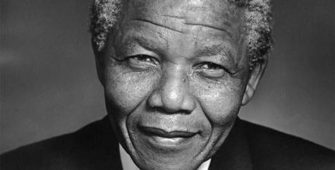A black and white photograph of Nelson Mandela wearing a black suit and leaning into the camera