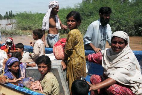 Many Pakistani children and one women sit in a boat filled with possessions on dirty flood waters whilst two men guide the boat from the outside