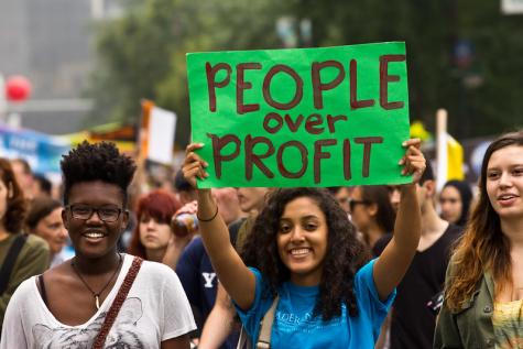 Protesters gather in the street. Three women are at the forefront of the image, they are smiling at the camera and hold a green poster which reads 'People over profit'