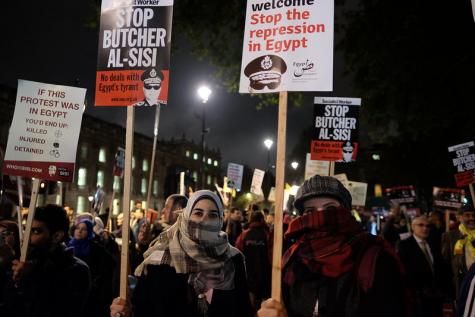 Many people stand in the cold night protesting against repression in Egypt holding banners and placards. Two women are in the foreground with their faces hidden.