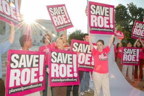 A group of people wearing pink tshirts stand holding posters saying Save Roe!