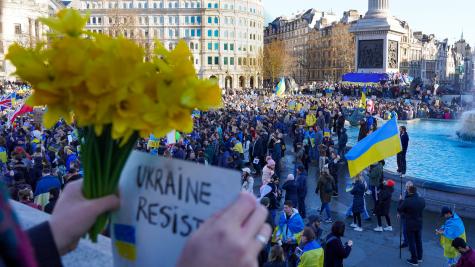 A scene of a pro-Ukraine protest in London's Trafalgar Square. Someone holds a bunch of yellow dafodils and a sign saying ' Ukraine resist' in the foreground. Below stands hundreds of protesters waving Ukraine flags and posters.