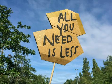 A wooden sign is being held up infront of some trees and a blue sky. It reads ' All you need is less'.