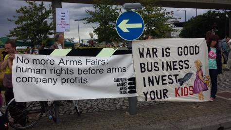 Two white banners are held up at a protest against the DSEi arms fair. They read 'human rights before arms company profits' and 'war is good business invest in your kids.'