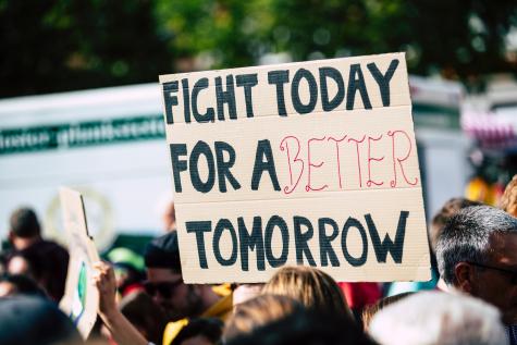 Protesters gather in the street, the main focus of the picture is a cardboard sign that one of the protesters is carrying, it reads 'Fight today for a better tomorrow.'