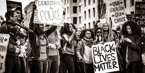 Black and white image of many protesters holding placards with calls to demilitarise the police. One young woman in the middle is yelling into a microphone.