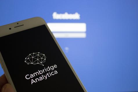 A phone displaying the logo of Cambridge Analytica is held up infront of a large screen showing the blue log in page for Facebook