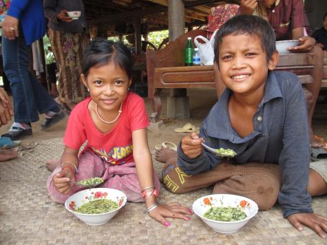 Two Cambodian children a girl and a boy sit on a woven grass carpet on the floor eating from bowls of enriched porridge there are various adults standing in the background