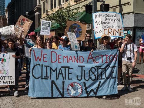 Global Climate Strike in Pittsburgh. Many protesters hold signs around a large white banner in the centre which reads 'We demand action. Climate justice now.'