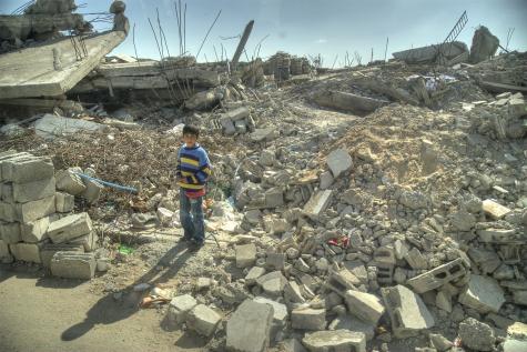 A young boy in Gaza stands in the middle of a huge pile of grey rubble from a destroyed building