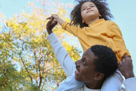 A smiling Black man wearing a white jumper carries a child on his shoulders, they are outside under a blue sky with a tree in the background