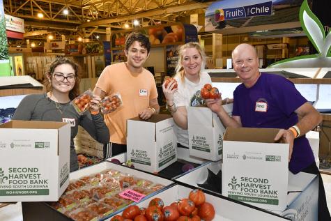 Two men and two women happily stand behind boxes of donated food in a food bank. They re wearing name tags and are holding fresh tomatos 