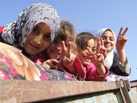 Four young girls from Gaza lean over a wall smiling and giving the peace sign to the camera