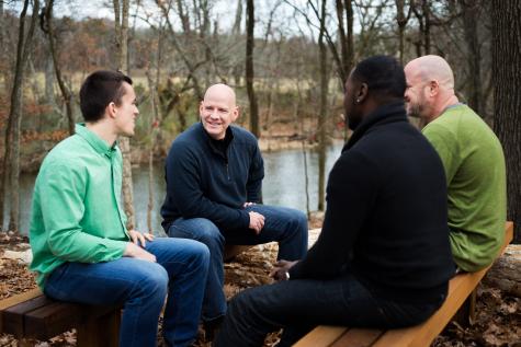 Four smiling men sit on two benches in the woods next to a river