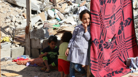 A girl and her two younger siblings stand in the rubble of a destroyed building in Gaza. There is a red sheet hanging to provide them with shade 