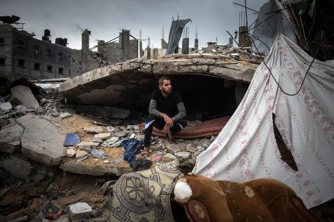 A man sits under a pile of broken infrastructure where he has a matress and a sheet hanging for privacy. The area is Khuzaa, the most destroyed part of the Gaza Strip