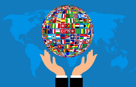 A large global filled with flags of the world is held by two white hands showing the white and black sleeves of his suit. The image sits infront of a blue background which is a map of the world