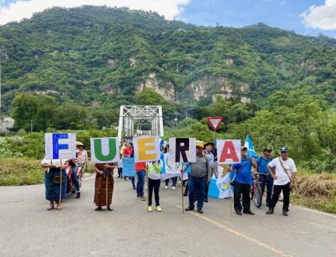 Five Guatemalan protesters stand infront of a bridge with a green mountain forming the back drop. They each hold a sign with a different letter spelling out the word 'fuera'.