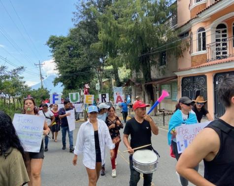 A group of protesters march down the street in Guatemala. They are carrying drums and whistles and horns to make noise. In the background there is a large sign that reads 'fuera' 