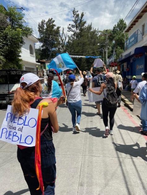 A shot from behind a group of people marching for democracy in Guatemala. Protesters have signs and are carrying flags 