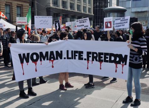 Three women from Iran hold a large white banner which says 'Woman, life, freedom' the slogan which became popular during the Mahsa Amini protests in Iran in 2022. Many protesters stand behind them.