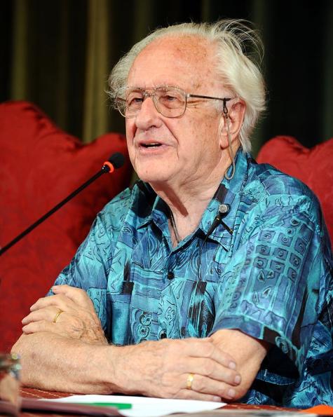Photograph of peace education pioneer Johan Galtung. He sits at a table with his arms crossed wearing a bright blue shirt, he has thinning grey hair and wears glasses.