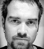 Black and white portrait of anti-war activist Ken O'Keefe. He is a white male with a short beard, short dark hair and two small tattoos on his face.