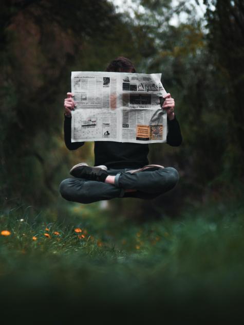 A man wearting dark clothes is crossed legged hovering above the forest floor holding up a newspaper