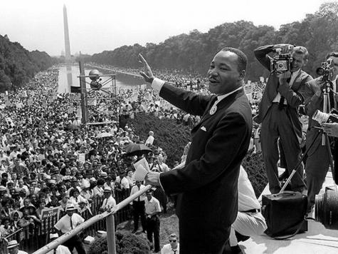 Black and white photo of civil rights leader Martin Luther King. He stands smiling wearing a suit on a stage infront of a large audience below. 