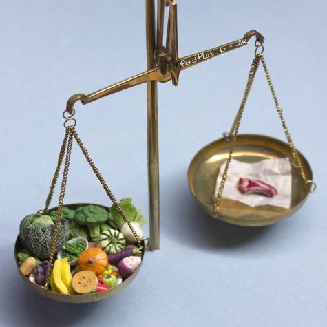 An old fashioned metal weighing scales shows one side filled with vegetables and the other side with just a small piece of meat in it. The scales are balanced. The image represents the huge environmental impact of meat in comparison with vegetables