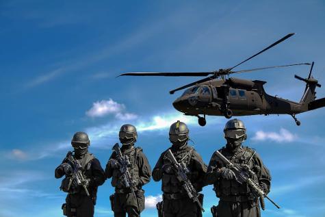 Four soldiers in full dark grey combat gear holding guns stand infront of a low flying helicopter. A blue sky fills the background.