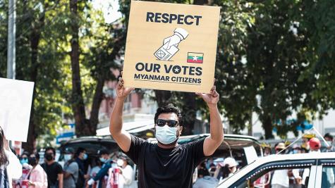 A male protester in Myanmar holds a large white sign which reads 'Respect our votes, Myanmar citizens' he is protesting again the military coup which took place in 2021. Many other protesters stand behind him.
