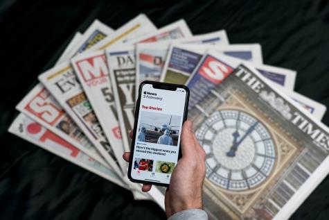 Nine British newspapers are spread out on a black surface while a mans hand holds a phone above them displaying a news site