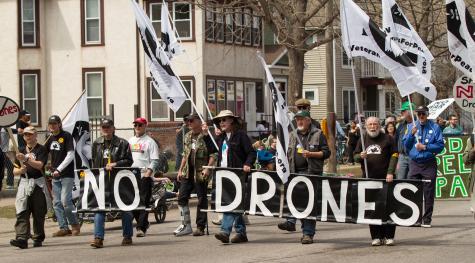 Veterans for Peace protesters walk down the street holding flags and a large banner which says ' No Drones.'