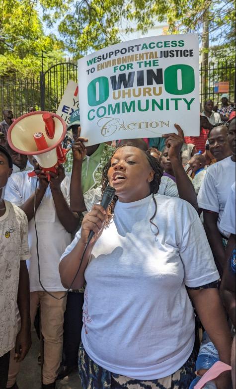 A female Kenyan activist holds a microphone at a protest. Other protesters have megaphones and there is a white poster which says ' We support access to environmental justice for the Uhuru community.