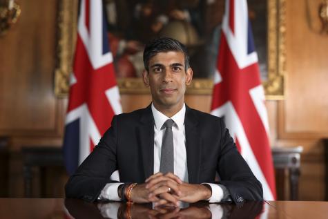 The British Prime Minister sits behind a wooden desk wearing a black suit with two red, white, and blue British flags on flag poles behind him. His hands are held together on the table and he smiles into the camera.