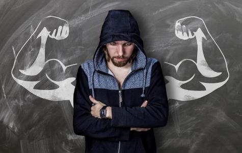 A sad looking man wearing a blue hoodie looks down at the floor with his arms crossed. He is standing infront of a chalkboard which has large curled biceps drawn on it