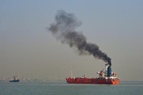 A large red cargo ship sits of the coastline spewing out large black clouds of pollution