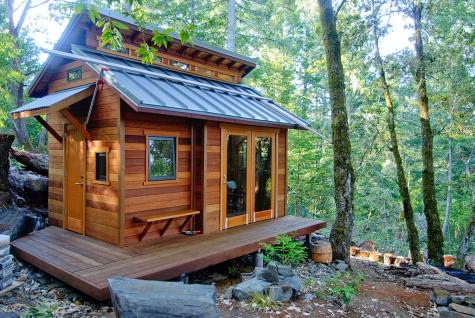 An eco-friendly wooden cabin tiny home sits in the middle of nature it has wooden decking around 2 sides and is surrounded by tress