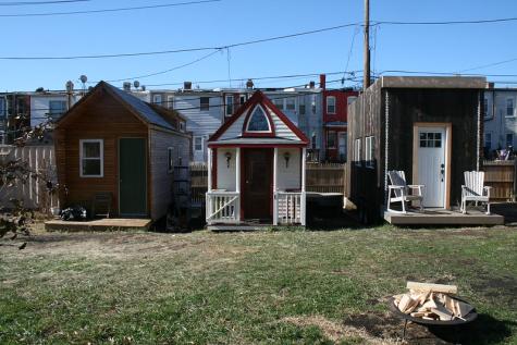 Three tiny homes stand side by side in Washington, DC. Two are brown, and the middle home is white and red. All three have small porches. There is a large grassy shared area infront of them with a fire pit in the foreground on the righthand side.