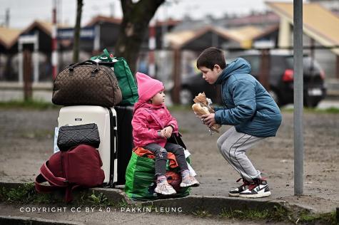 A young Ukranian boy hands his little sister a sandwich. She is sitting on a bag on the floor infront of many suitcases.