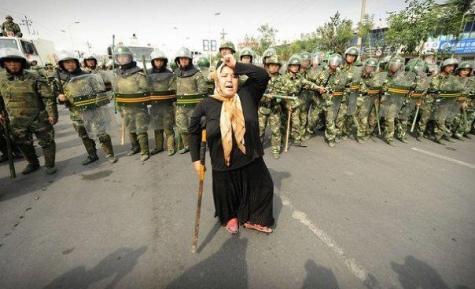 An older Uyghur lady wearing a yellow headscarf stands using a crutch and holding her hand against her head in anguish. Behind her is a line of Chinese soldiers wearing full riot gear.