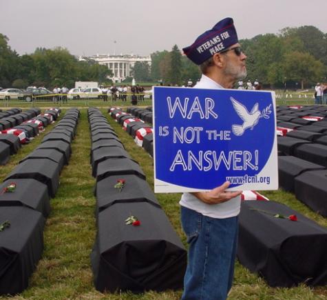 A man wearing a Veterans for Peace hat stands infront of war graves holding a blue sign that says 'War is not the answer.'