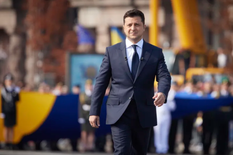 Volodymyr Zelenskyy, the President of Ukraine walks through the Parade of Troops in a blue black suit. He looks serious and aways from the camera, everything in the background has been blurred
