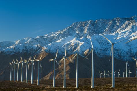 Two rows of white wind turbines are shown infront of a beautiful miuntain scene. The top half of the mountains are covered in snow