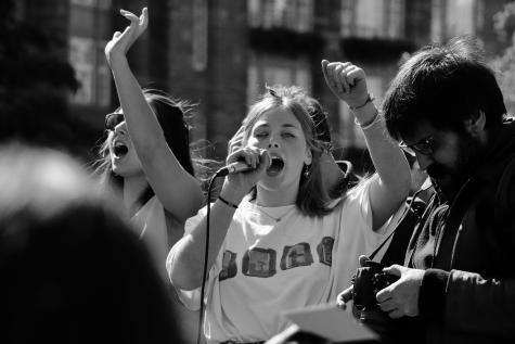 A young girl stands in a crowd with her fist raised. She is shouting into a microphone