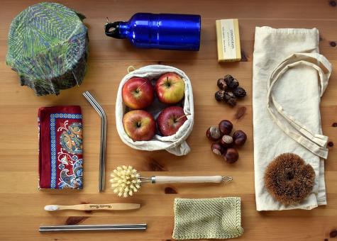 Many items that allow people to live without creating waste sit on a wooden table. There is a tote bag, reusable packaging, a bamboo toothbrush, a water bottle, some natural soap, and reusable metal straws